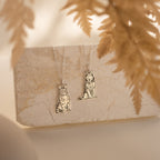 Two Custom Pet Portrait Necklaces, each with a cat and dog pendant, are displayed on a marble slab with dried leaves in the foreground.