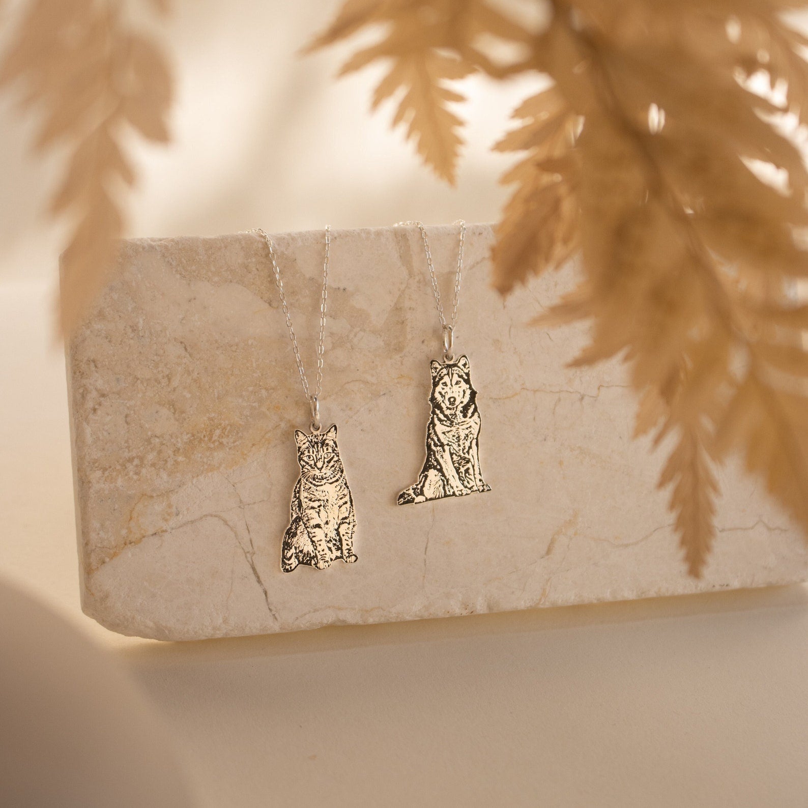 Two Custom Pet Portrait Necklaces, each with a cat and dog pendant, are displayed on a marble slab with dried leaves in the foreground.