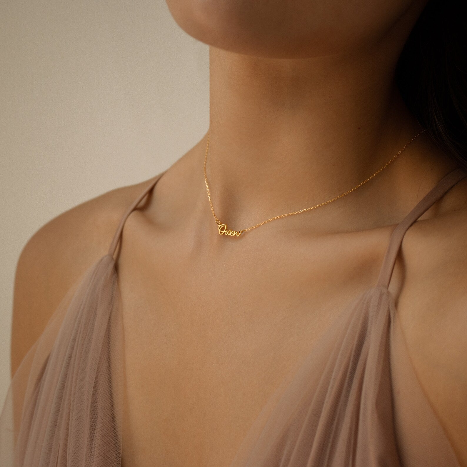 A woman wears the Tiny Grace Name Necklace with a cursive pendant, paired with a sheer taupe spaghetti strap top.