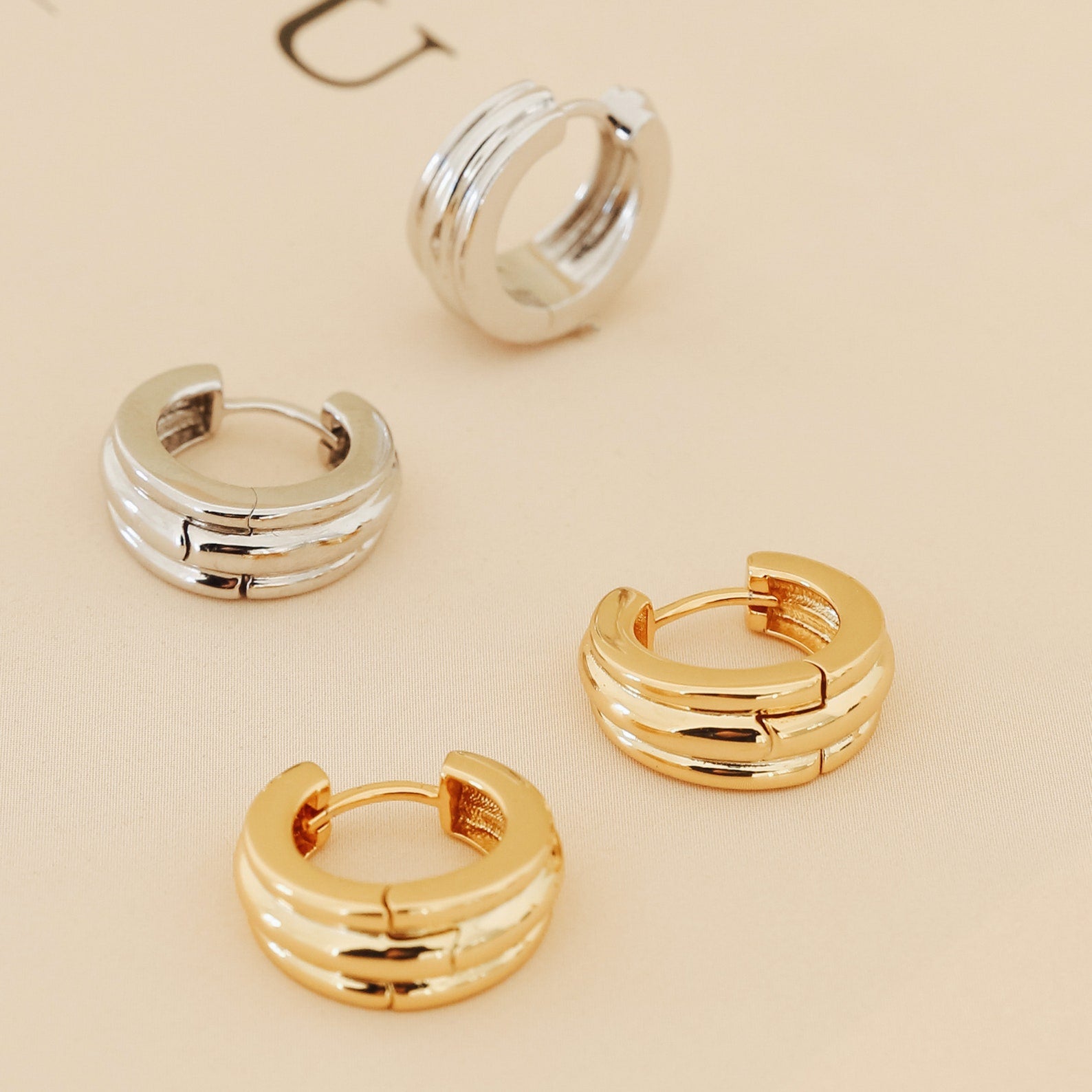 Two pairs of Canal Chunky Hoops—one silver, one gold—are displayed on a beige surface. These contemporary earrings are perfect for everyday wear and add a modern touch to any look.