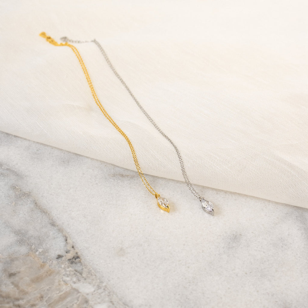 The Athens Diamond Necklace, a marquise diamond pendant on a dainty cable chain in gold or silver, is elegantly displayed on marble and fabric surfaces.