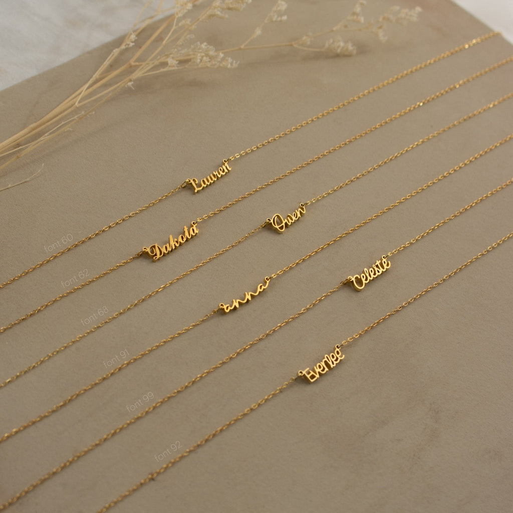 Six gold personalized name necklaces, including the Tiny Grace Name Necklace, are arranged on a beige surface with dried flowers placed above them.