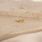 The Trio Sphere Studs feature a small gold three-dot cluster design, displayed on a beige stone surface—ideal for creating an elegant earring stack or accenting alternative piercings.