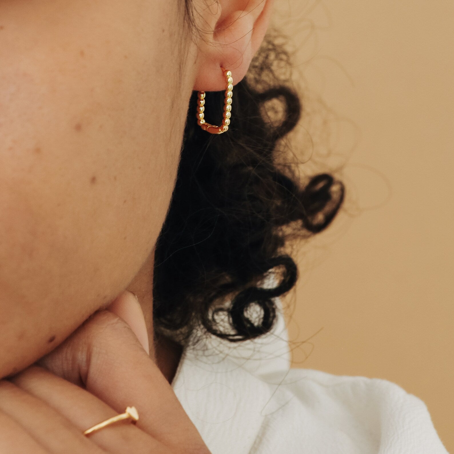 A close-up of a woman with curly hair wearing the chic Indiana Beaded Hoops and a gold ring, set against a beige background.