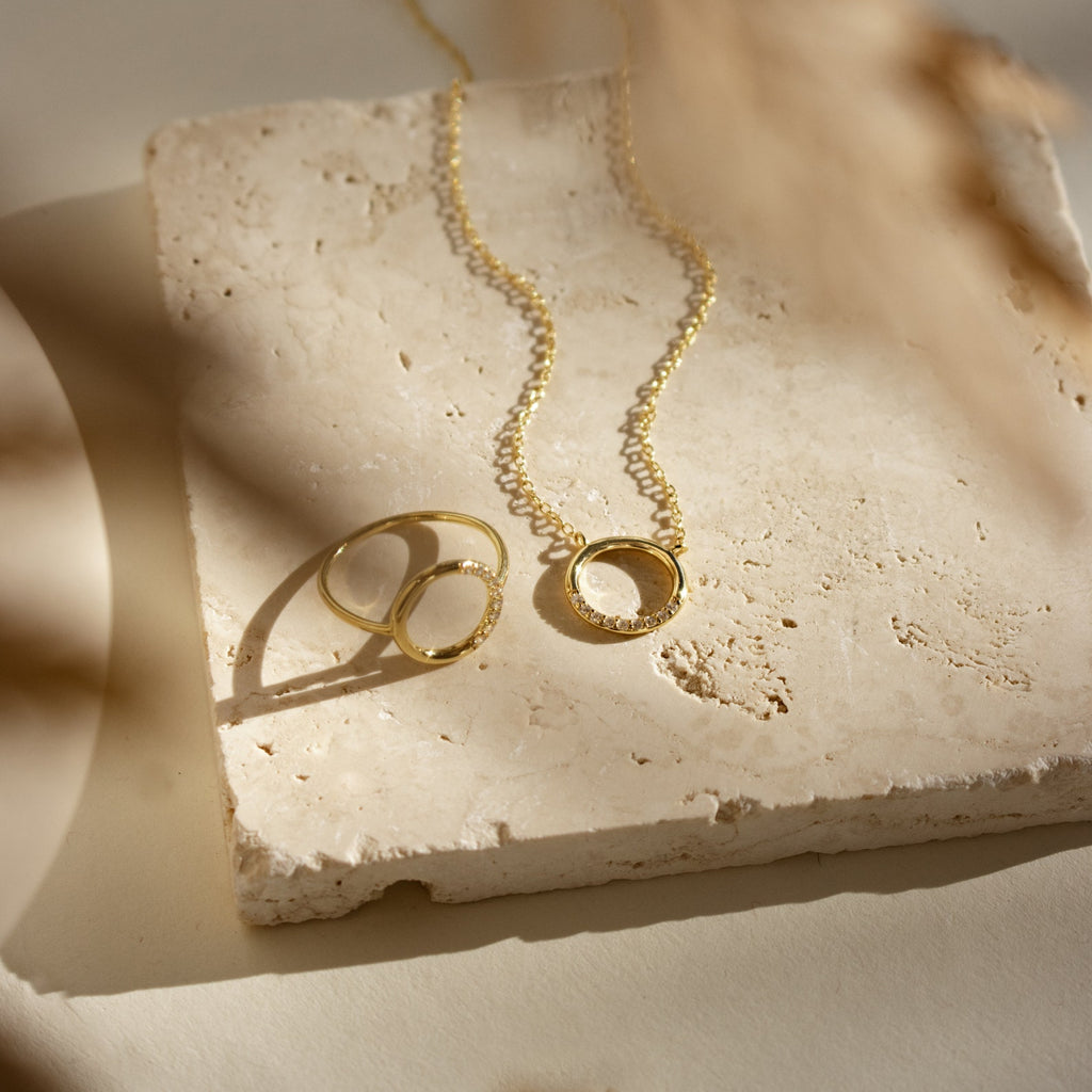 The Kayla Pave Circle Necklace, featuring a gold round pendant, rests on a textured stone slab with a soft shadow in warm light—a minimalistic jewelry piece ideal for any occasion.