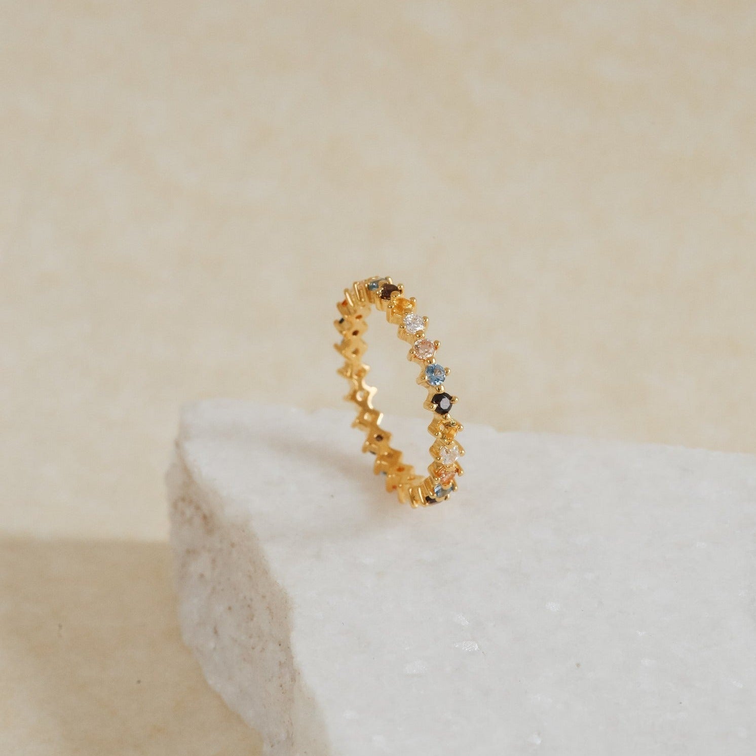 The Rainbow Crystal Ring, featuring pastel-toned gemstones set in delicate gold, rests on a textured white stone surface.