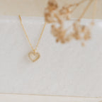 The Forever Pave Heart Necklace features a gold heart pavé design with small crystals, ideal for bridal jewelry, elegantly displayed on white marble with dried flowers.