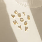 Gold alphabet charms spelling CUTE and AMOYBK on a sunlit surface—our Dangling Pave Initial Necklace is a delicate, sparkling piece, perfect as a necklace or thoughtful gift for women.