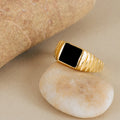 The Black Signet Ring, featuring a black rectangular stone set in gold, is displayed on a smooth beige pebble near a larger rock.