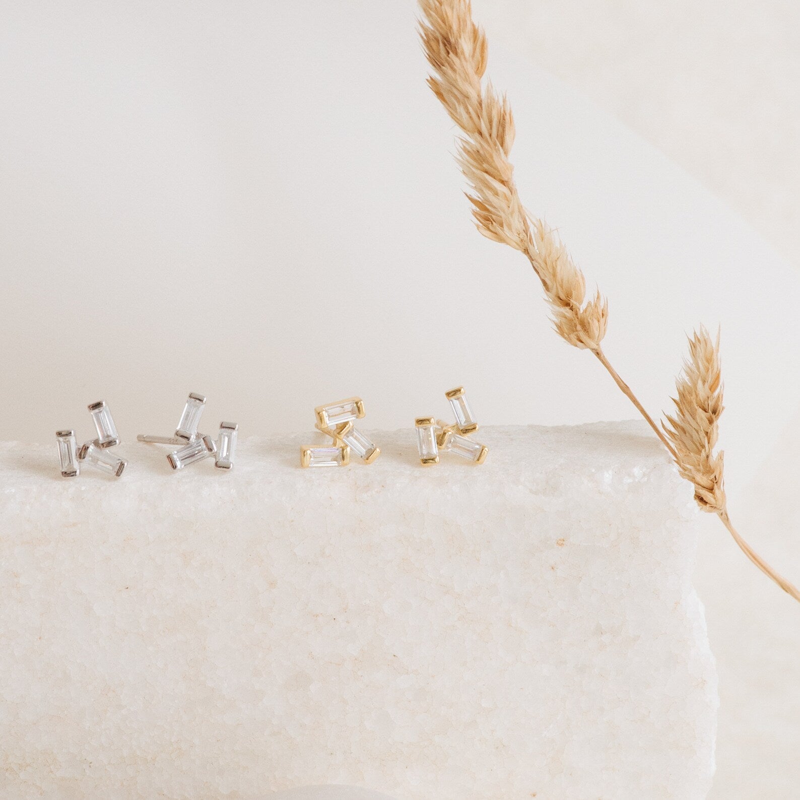 The Glacier Diamond Studs set includes two pairs of stud earrings—one silver and one gold with elegant baguette stones—displayed on a white stone beside a dried wheat stalk.