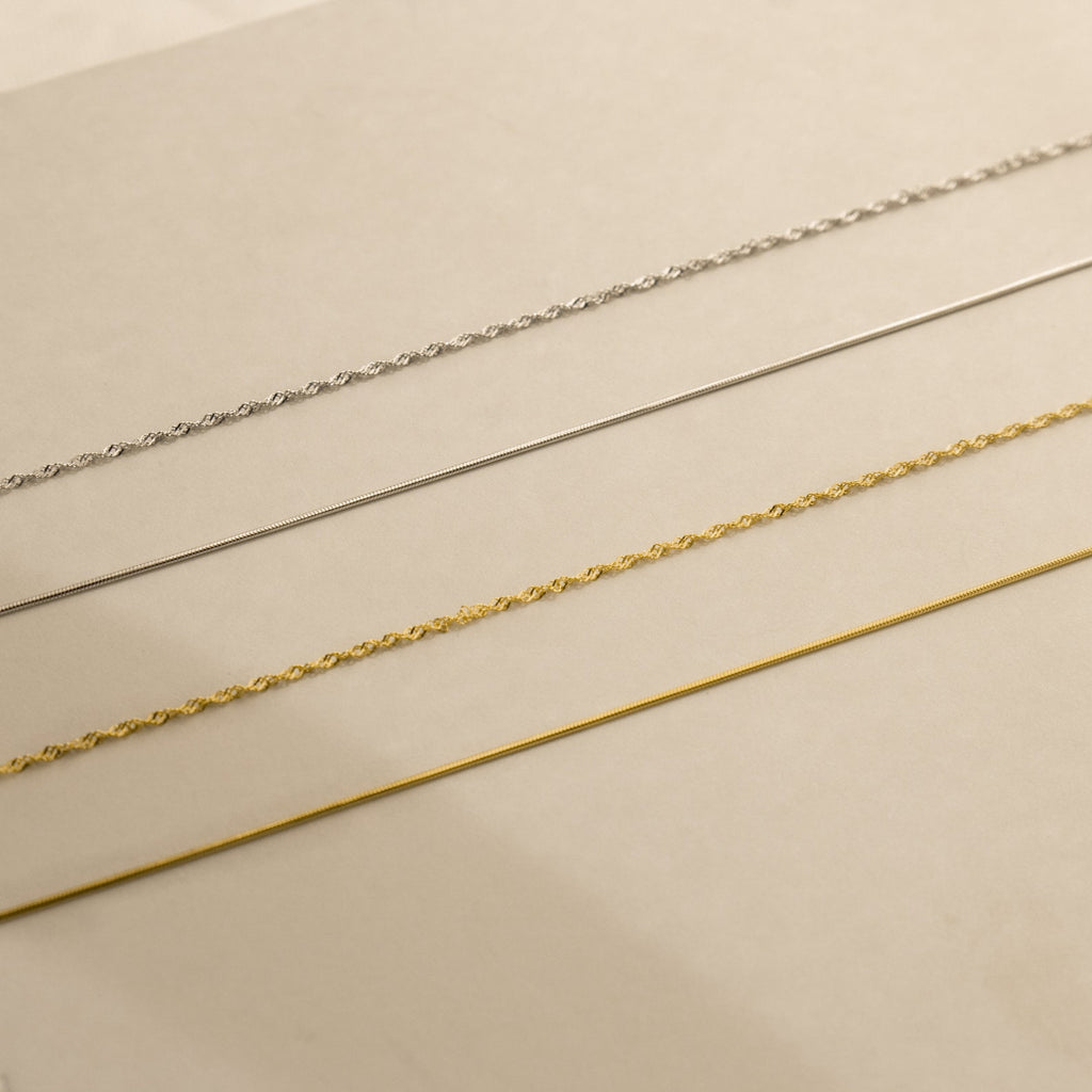 Four delicate necklaces—including the Monet Duo Chain—are arranged in parallel lines on a beige surface.