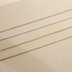 Four delicate necklaces—including the Monet Duo Chain—are arranged in parallel lines on a beige surface.