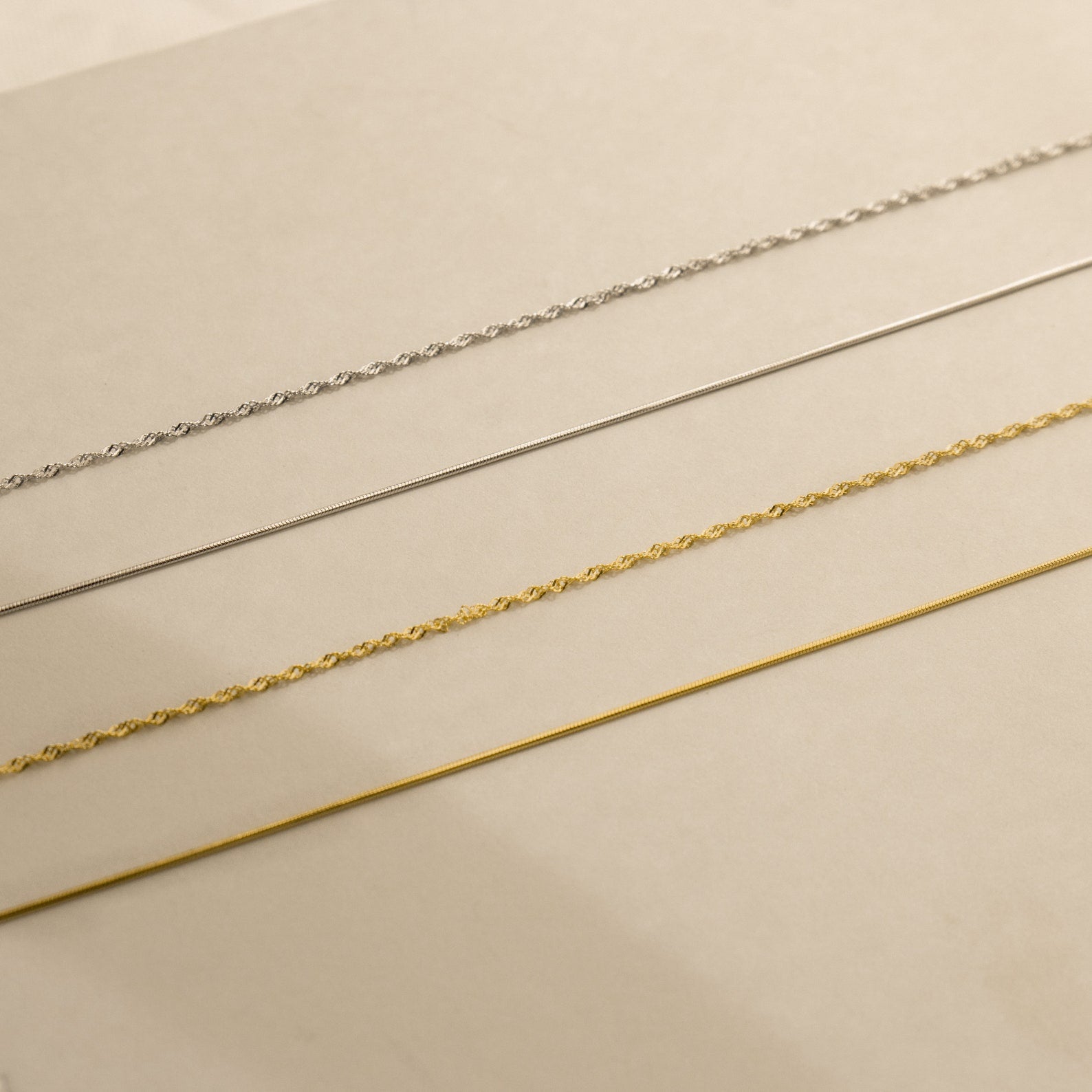 Four delicate necklaces—including the Monet Duo Chain—are arranged in parallel lines on a beige surface.