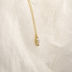 The Athens Diamond Necklace features a gold chain with a clear marquise diamond pendant, shown on a soft cream-colored fabric background.