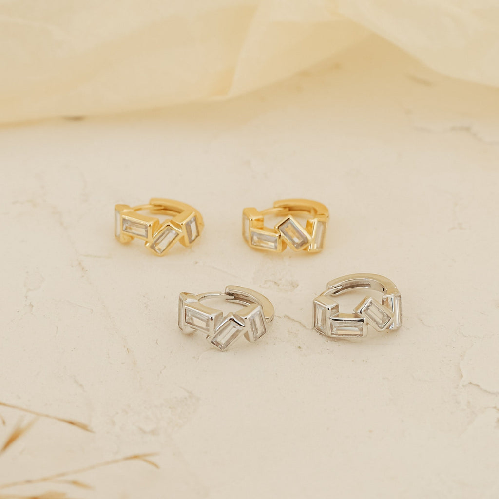 The Glacier Baguette Huggies, featuring two gold and two silver ear cuffs with a minimalistic sparkle, are displayed on a cream-colored textured surface.
