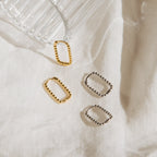 Four stylish Indiana Beaded Hoops—two gold and two silver—are displayed on white fabric by a glass cup, bringing a modern accent to your accessory collection.