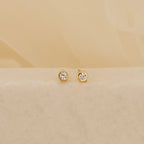The Jane Diamond Studs are gold earrings with round brilliant diamonds, shown on a light beige surface against a soft background.