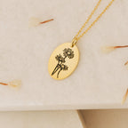 The Oval Flower Necklace features an engraved gold oval pendant with three flowers, displayed on a light textured surface with dried petals—an elegant birth flower necklace, perfect for unique flower jewelry lovers.
