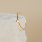 The Nala Tiny Heart Ring, featuring a small heart detail, rests on a textured white surface with a beige background.