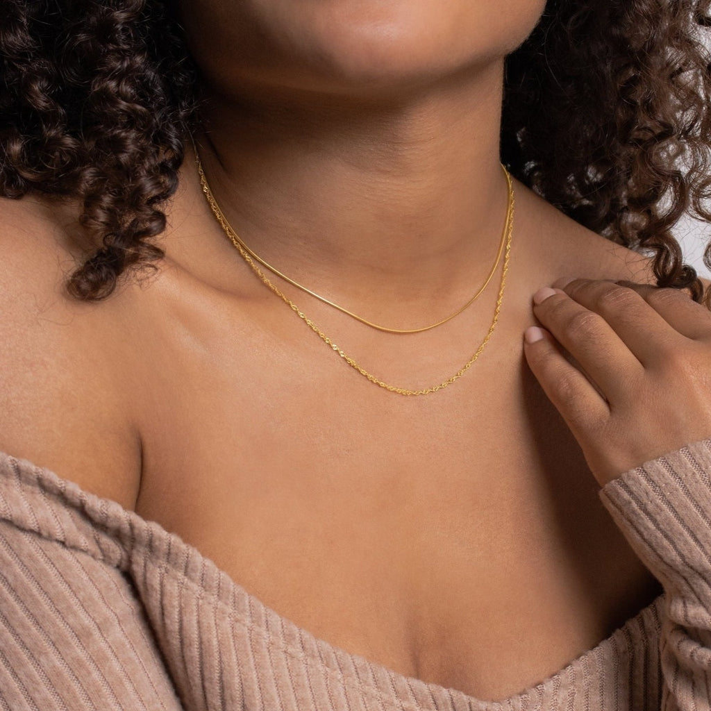 Close-up of a woman wearing two layered gold necklaces: a sleek, flat snake chain and a faceted bead-style chain, styled with a ribbed off-the-shoulder top.