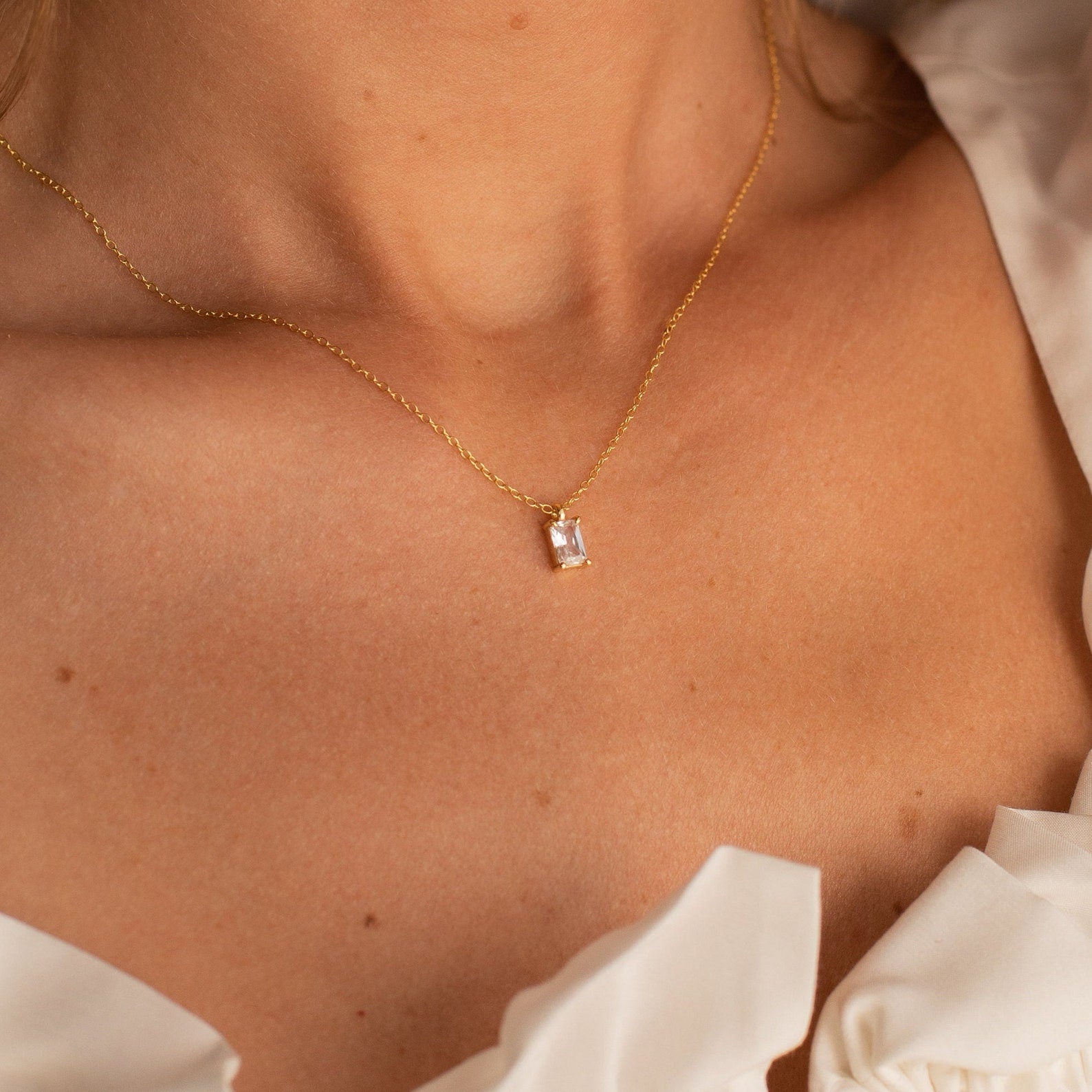 Gold necklace featuring a rectangular emerald-cut crystal pendant, worn by a model in a white ruffled top, highlighting the clean, geometric lines and radiant clarity of the stone.