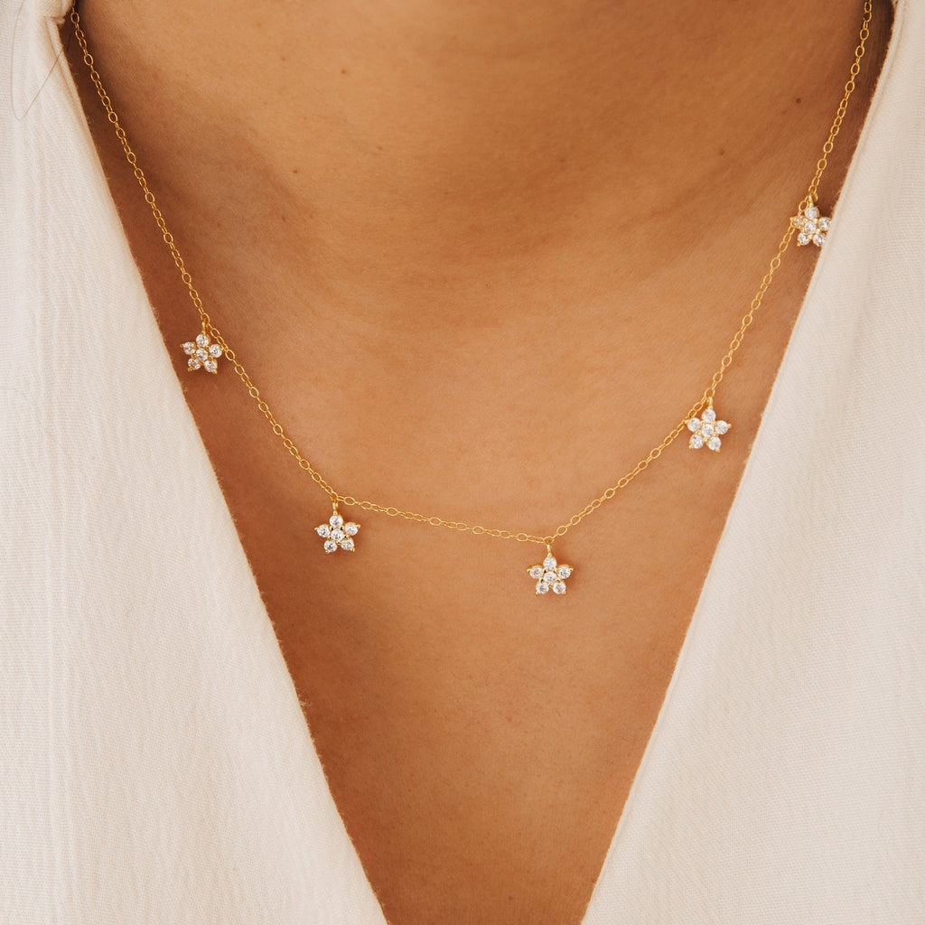 The Nanala Diamond Flower Necklace, featuring delicate flower-shaped charms, sparkles on a person in a white top, bringing an elegant touch to any outfit.