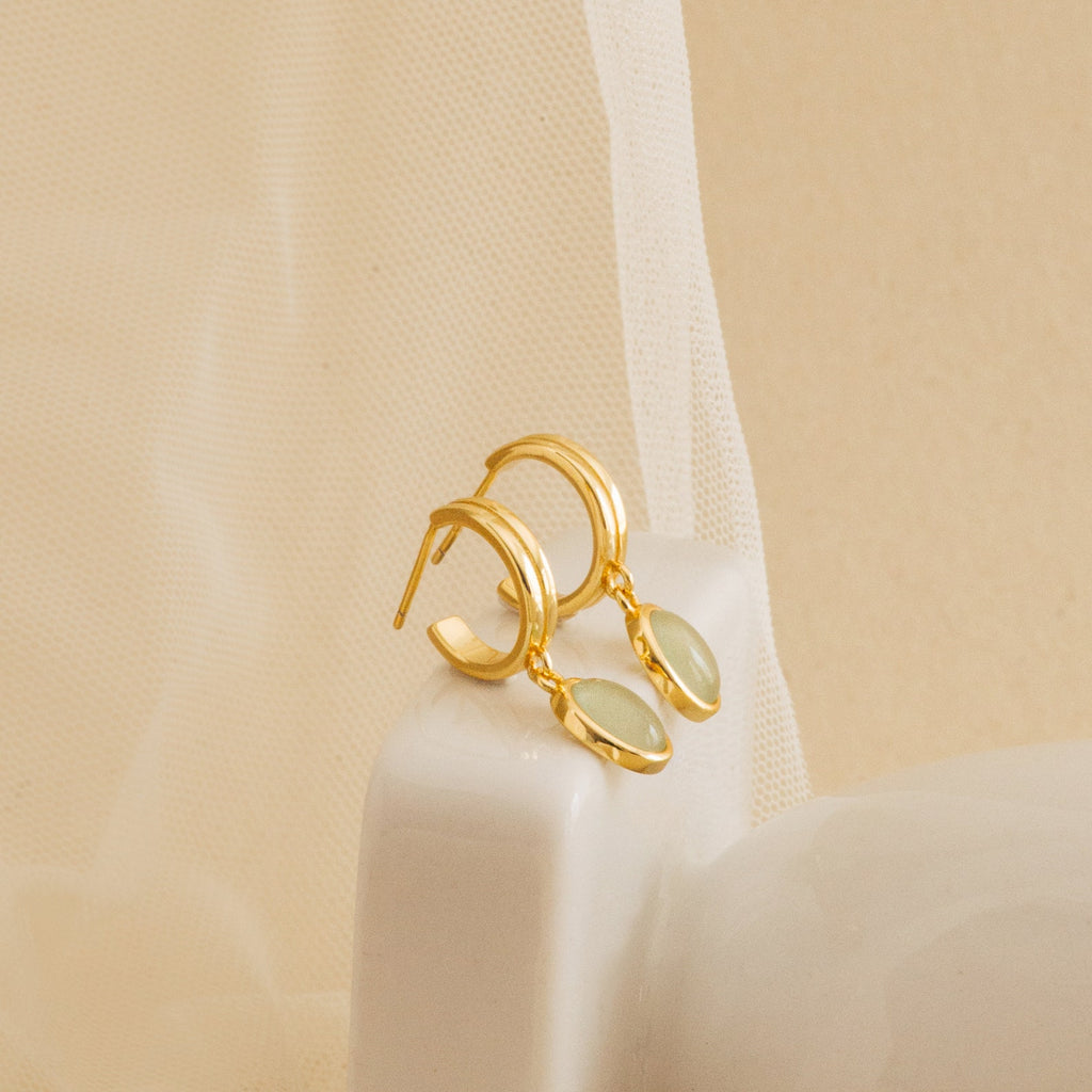 Gold huggie hoop earrings with oval jade gemstone charms displayed on a soft mesh fabric, highlighting their refined and minimalist aesthetic.