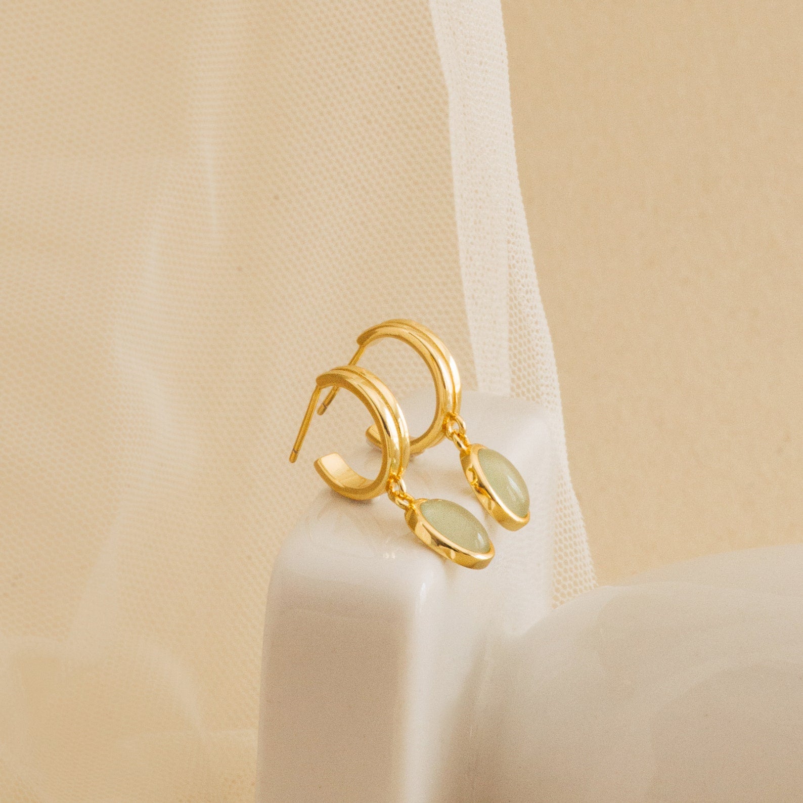 Gold huggie hoop earrings with oval jade gemstone charms displayed on a soft mesh fabric, highlighting their refined and minimalist aesthetic.