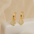 Pair of gold huggie hoop earrings with oval jade gemstone drop charms displayed on a soft neutral fabric, showcasing a dainty and timeless design.