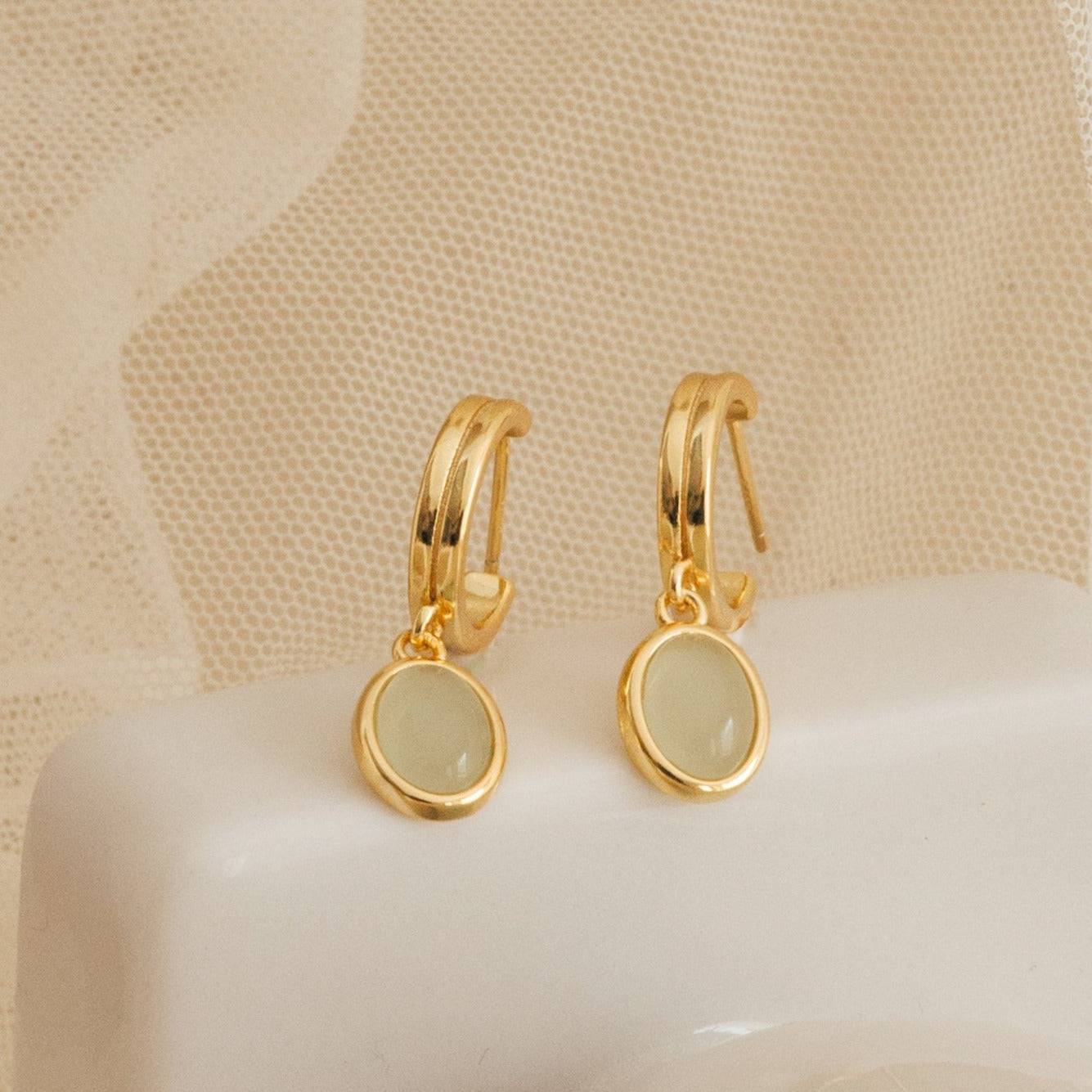 Pair of gold huggie hoop earrings with oval jade gemstone drop charms displayed on a soft neutral fabric, showcasing a dainty and timeless design.