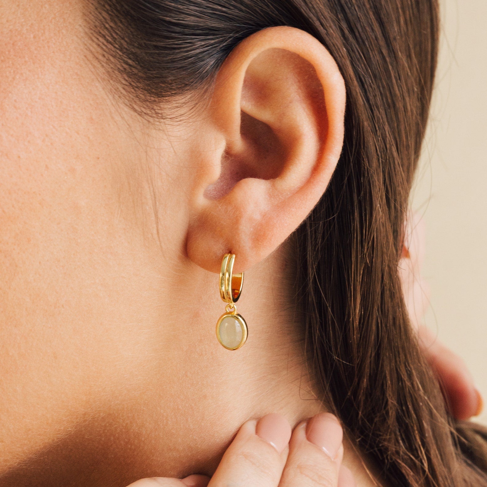 Close up of a model wearing gold huggie hoop earrings with a dangling oval green jade gemstone charm, styled as elegant minimalist everyday jewelry.
