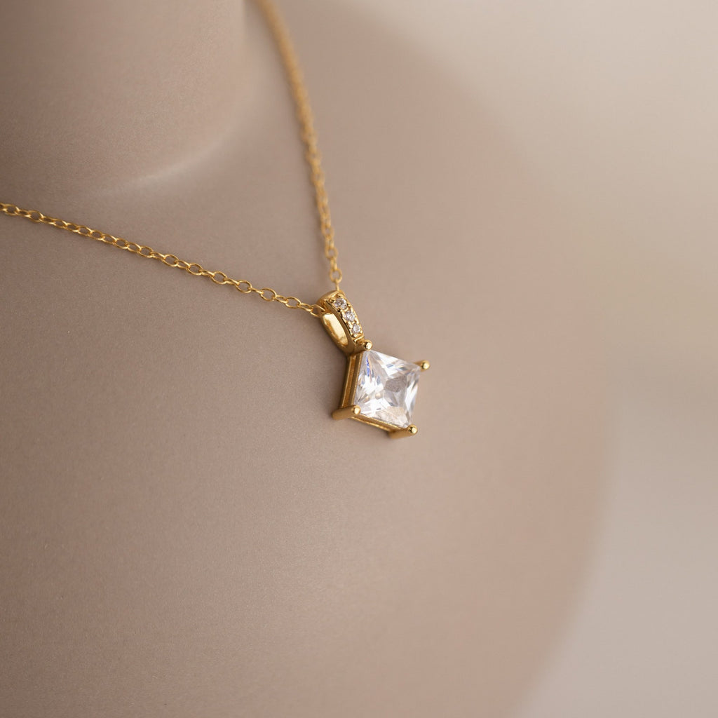 Close-up of a gold necklace featuring a square-cut clear gemstone pendant secured by four prongs, with tiny accent stones lining the bail, displayed on a neutral-toned bust.