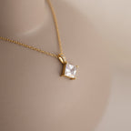 Close-up of a gold necklace featuring a square-cut clear gemstone pendant secured by four prongs, with tiny accent stones lining the bail, displayed on a neutral-toned bust.