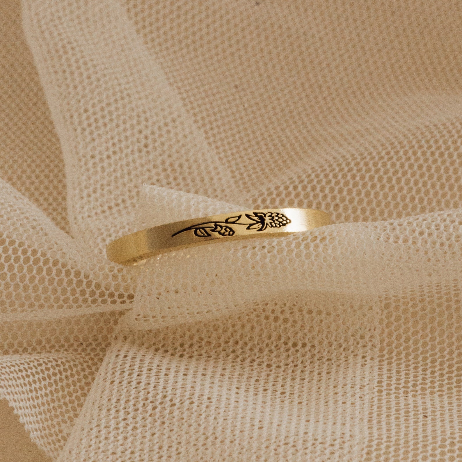 The Stacking Flower Band, featuring a delicate floral engraving, rests on soft beige mesh fabric, highlighting the elegance of botanical-inspired jewelry.