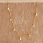 Delicate gold chain necklace with small dangling freshwater pearls, displayed against a textured beige surface.