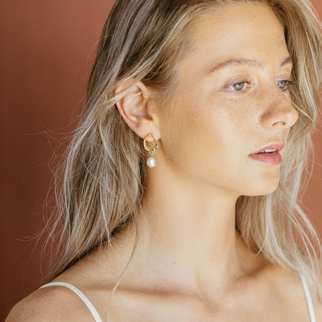 A woman with long blonde hair wears Rebecca Pearl Drop Earrings—elegant freshwater pearl drops—against a brown background.