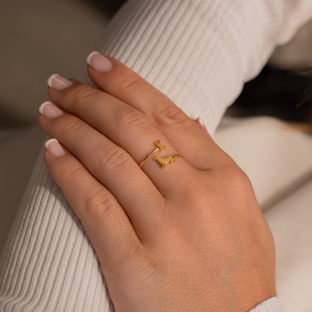 A hand with pale pink manicured nails wears the Duo Pet Initial Wrap Ring in Rose Gold, designed with two cat shapes, on the ring finger.