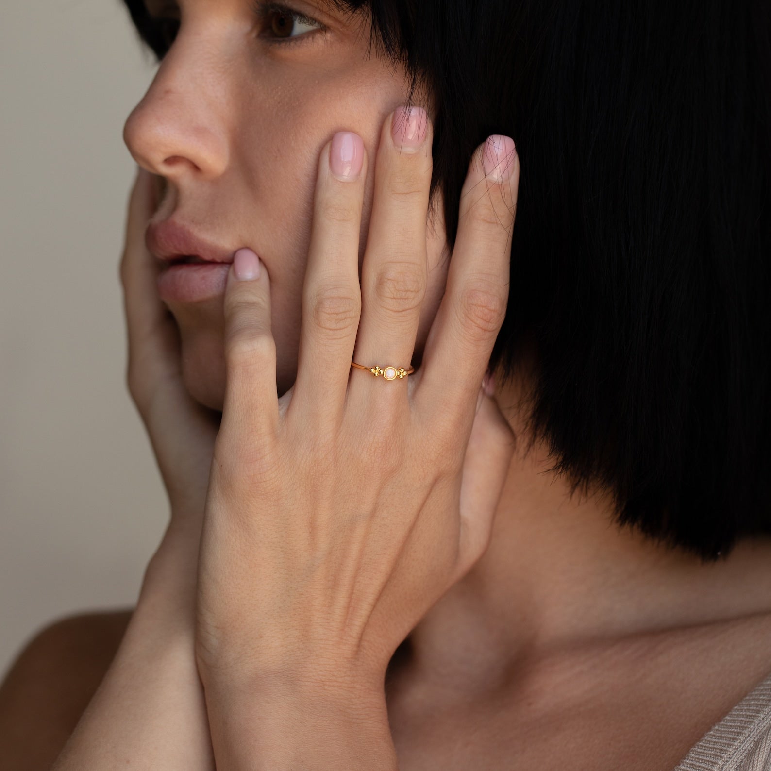 A woman with short black hair touches her face, showcasing the Sophia Opal Ring—a vintage-inspired piece adorned with pearls and an opal stone.