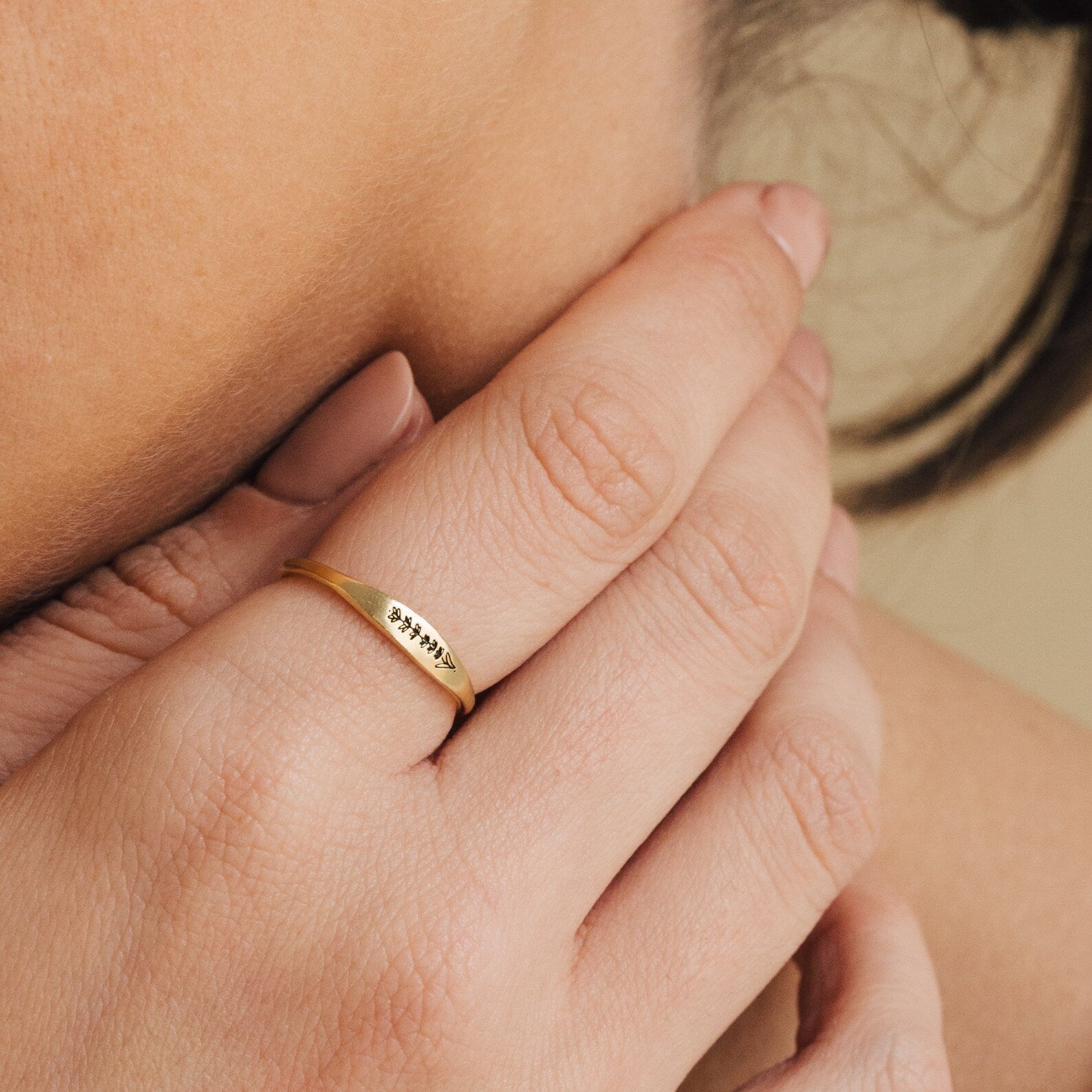 A hand with nude nails wears the Skinny Flower Ring in Rose Gold, featuring a delicate engraved leaf pattern, gently touching the neck.