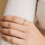 A hand with nude nails wears the Flower Bar Ring in Rose Gold, a delicate skinny band with leaf engravings, resting on a white textured sleeve—an elegant piece of botanical jewelry.