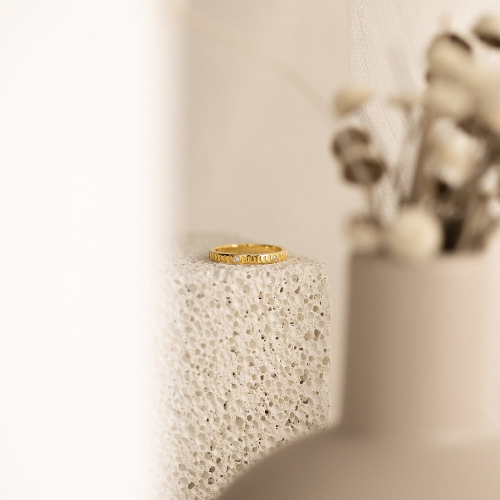 The Augustine Moon Phase Ring rests on a textured stone surface, with blurred dried flowers in the foreground.
