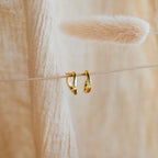 The McKenna Abstract Hoops feature an organic gold design, displayed against soft beige fabric and dried grass—perfect for elevating your everyday look with a touch of unique elegance.
