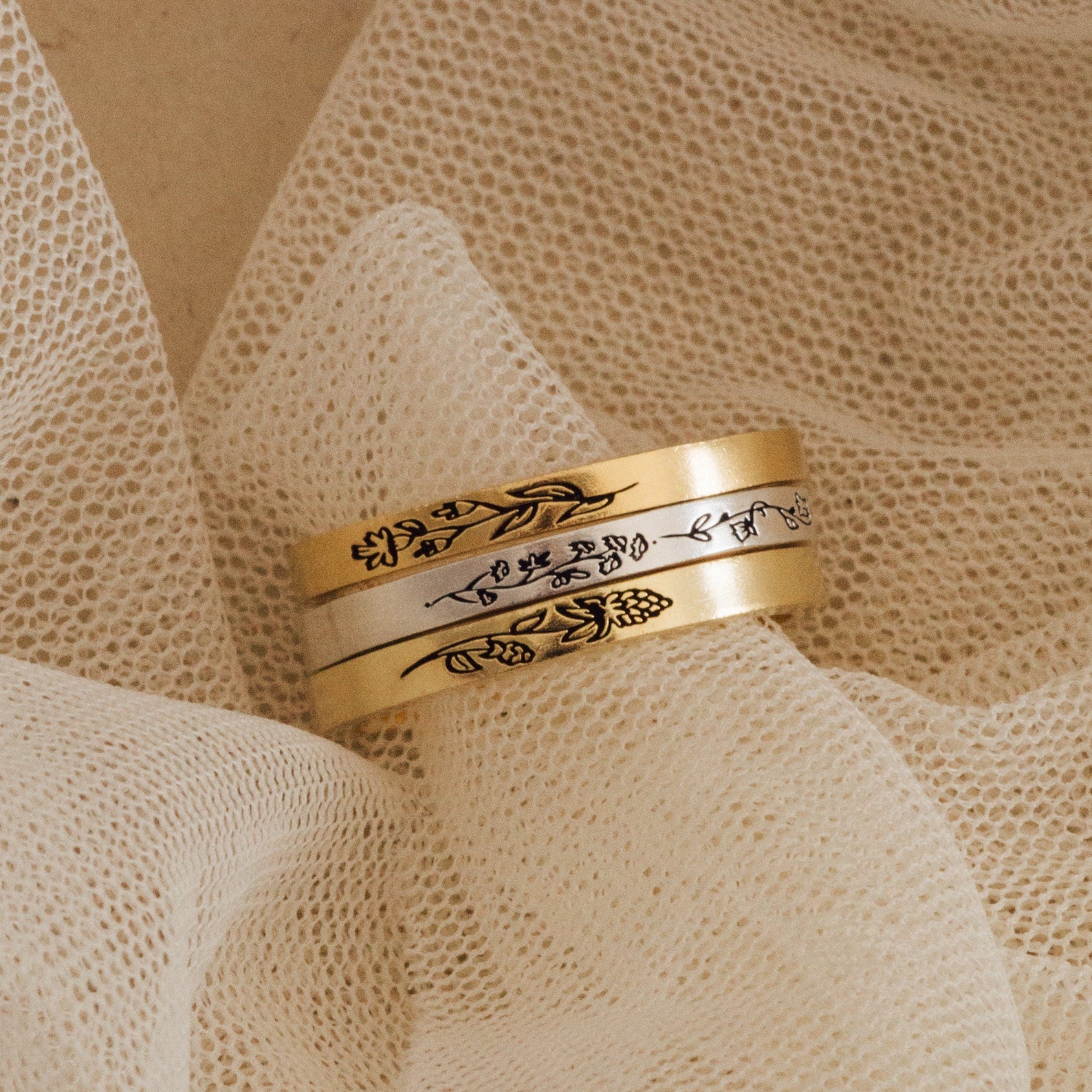The Stacking Flower Band, an engraved gold and silver ring with a floral design, rests on textured cream fabric—an ideal piece for lovers of botanical jewelry.