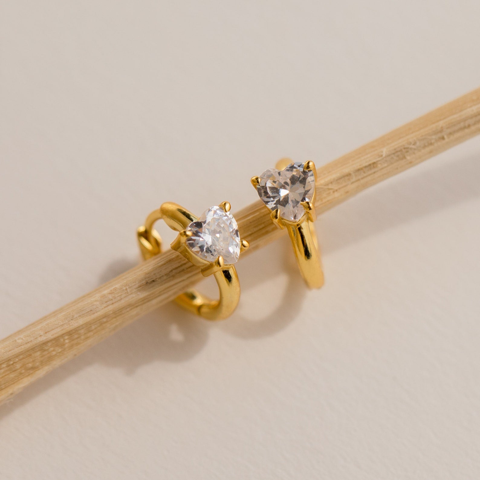 Showcased on a thin beige stick, the Diamond Heart Huggies feature two gold rings with heart-shaped clear stones—dainty, hoop-inspired earrings that make a perfect gift against a neutral backdrop.