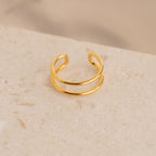 The Everly Thin Duo Band is a gold open cuff ring featuring an adjustable double band design, ideal for stacking. Shown on a textured beige stone surface.