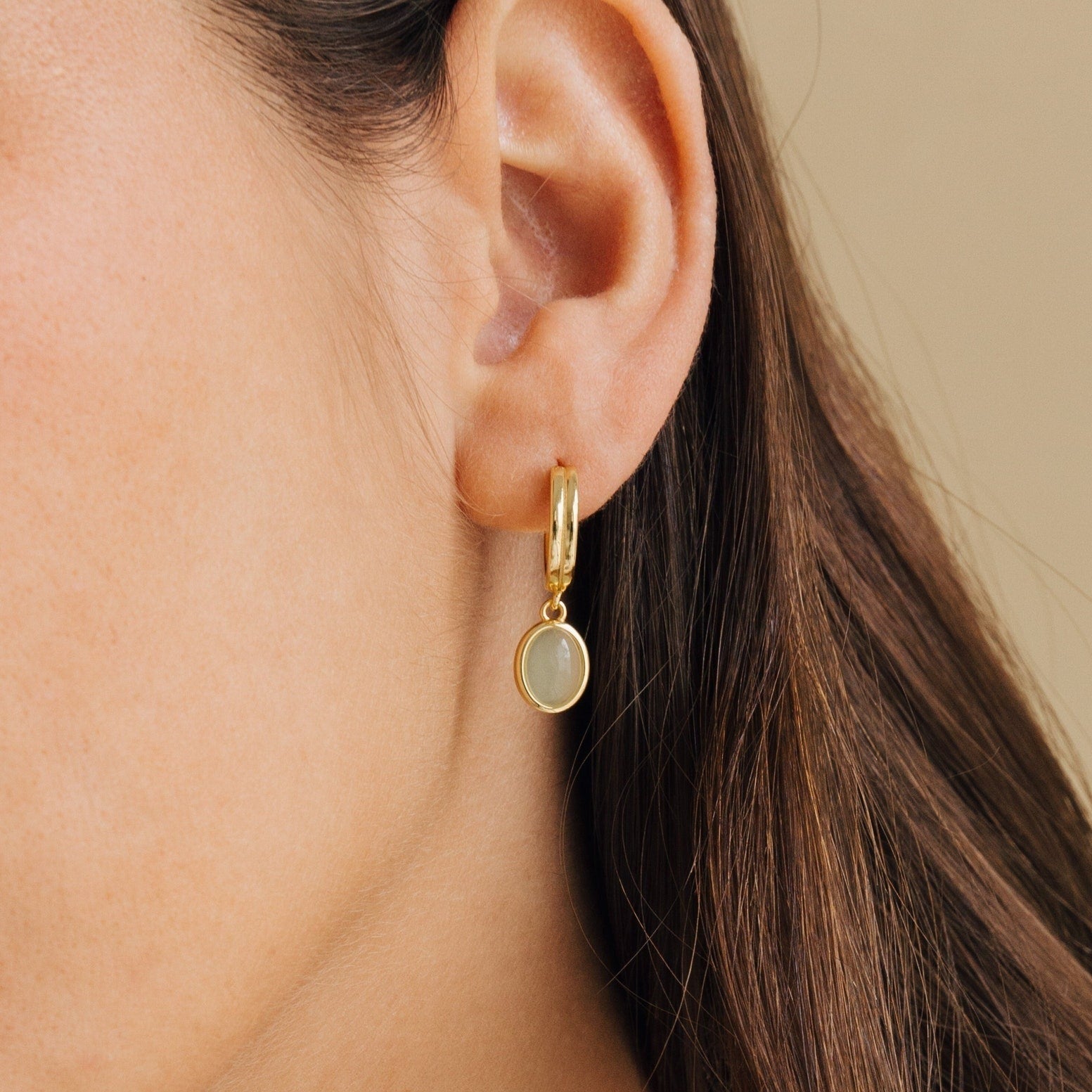 Side profile of a model wearing gold hoop earrings with a small oval jade gemstone pendant, styled as delicate everyday jewelry with a natural look.