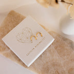 The Tiny Circle & Triangle Huggies—lightweight gold minimalist earrings—are shown on a textured white jewelry box with a geometric heart and Caitlyn Minimalist text, adding chic style to any earring stack.