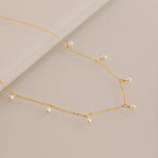 Close up of a delicate gold chain necklace with evenly spaced small white pearl drops, displayed on a smooth beige surface.