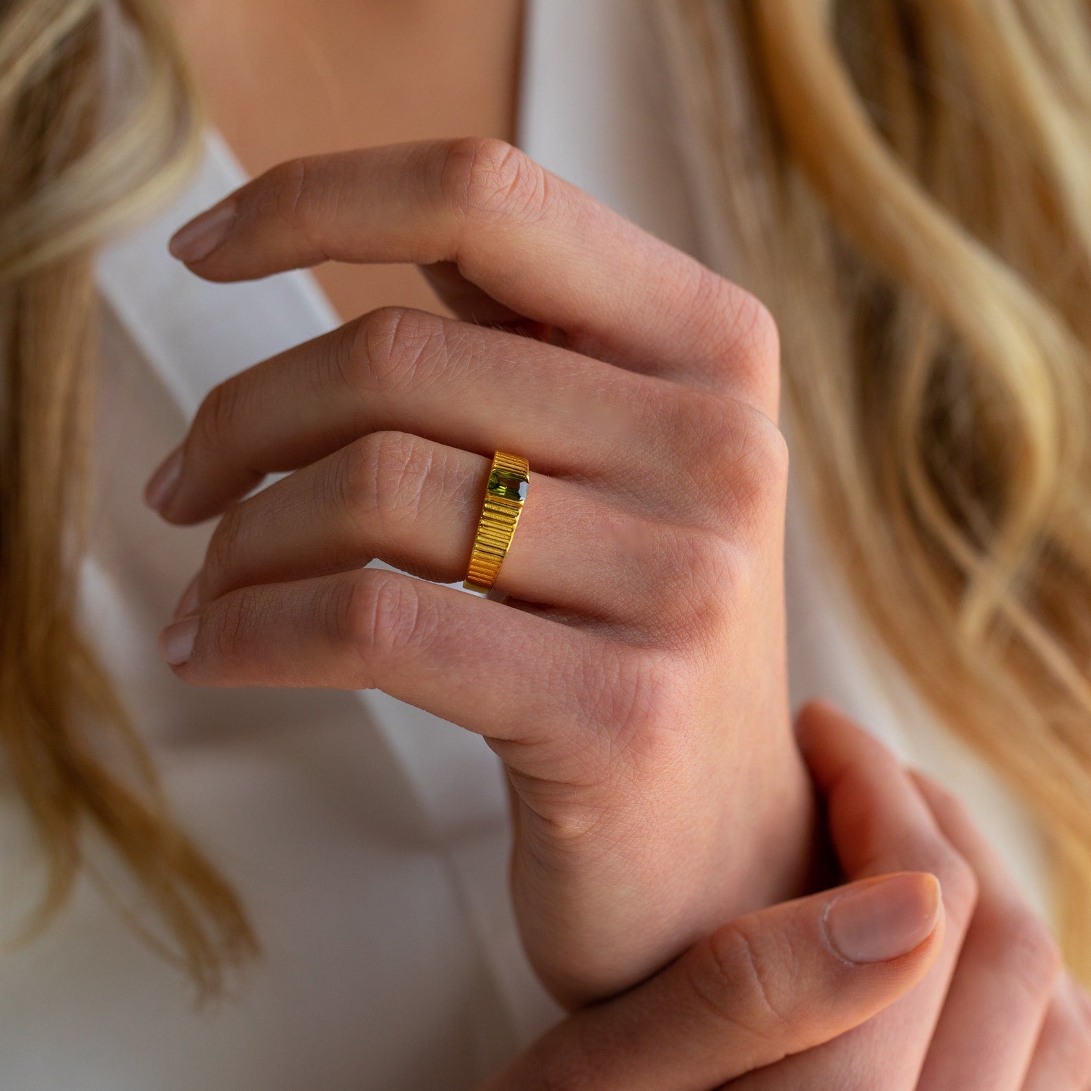 A hand adorned with the Olive Peridot Signet Ring, featuring a small green gemstone, fingers gently touching against a softly blurred background.