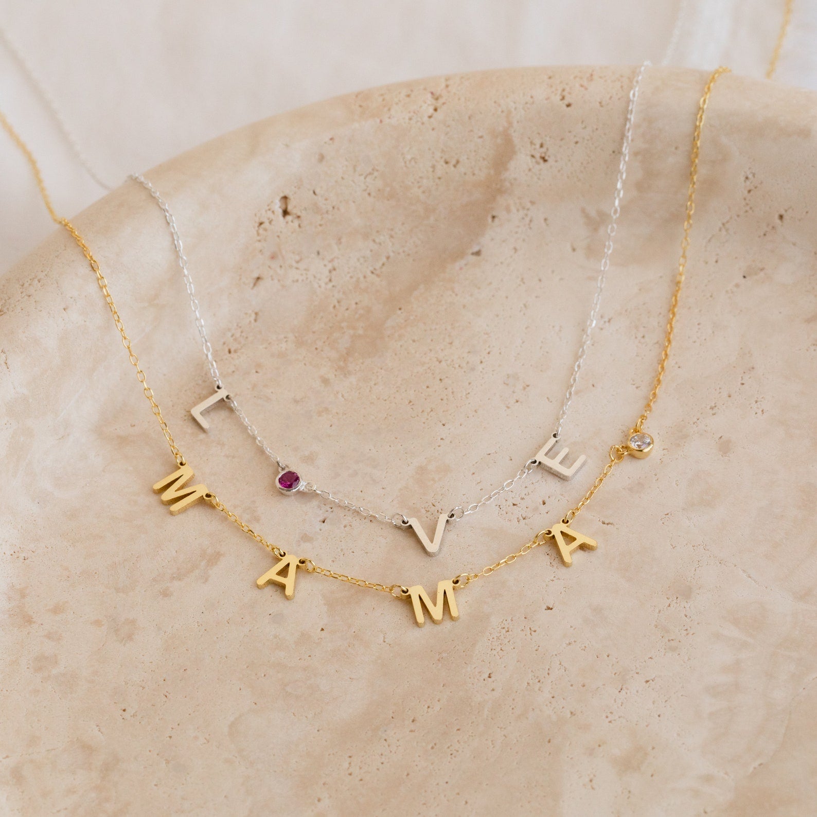 The Alice Letter Birthstone Necklace spells LOVE or MAMA with gold charms, set on a beige stone plate—a beautiful personalized gift for mom.